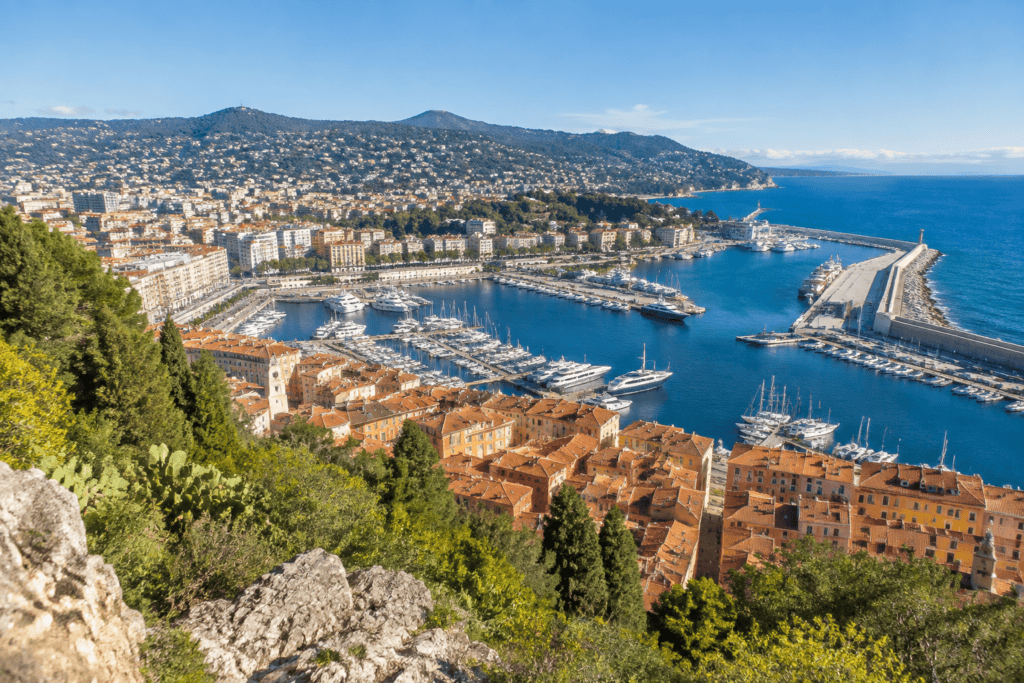 Panorama du port de Nice et des toits ocre du Vieux-Nice, encadré de végétation et de rochers au premier plan.