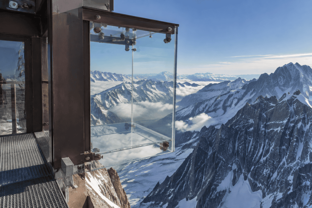 Une boîte vitrée fixée à une structure métallique surplombe des sommets alpins enneigés.