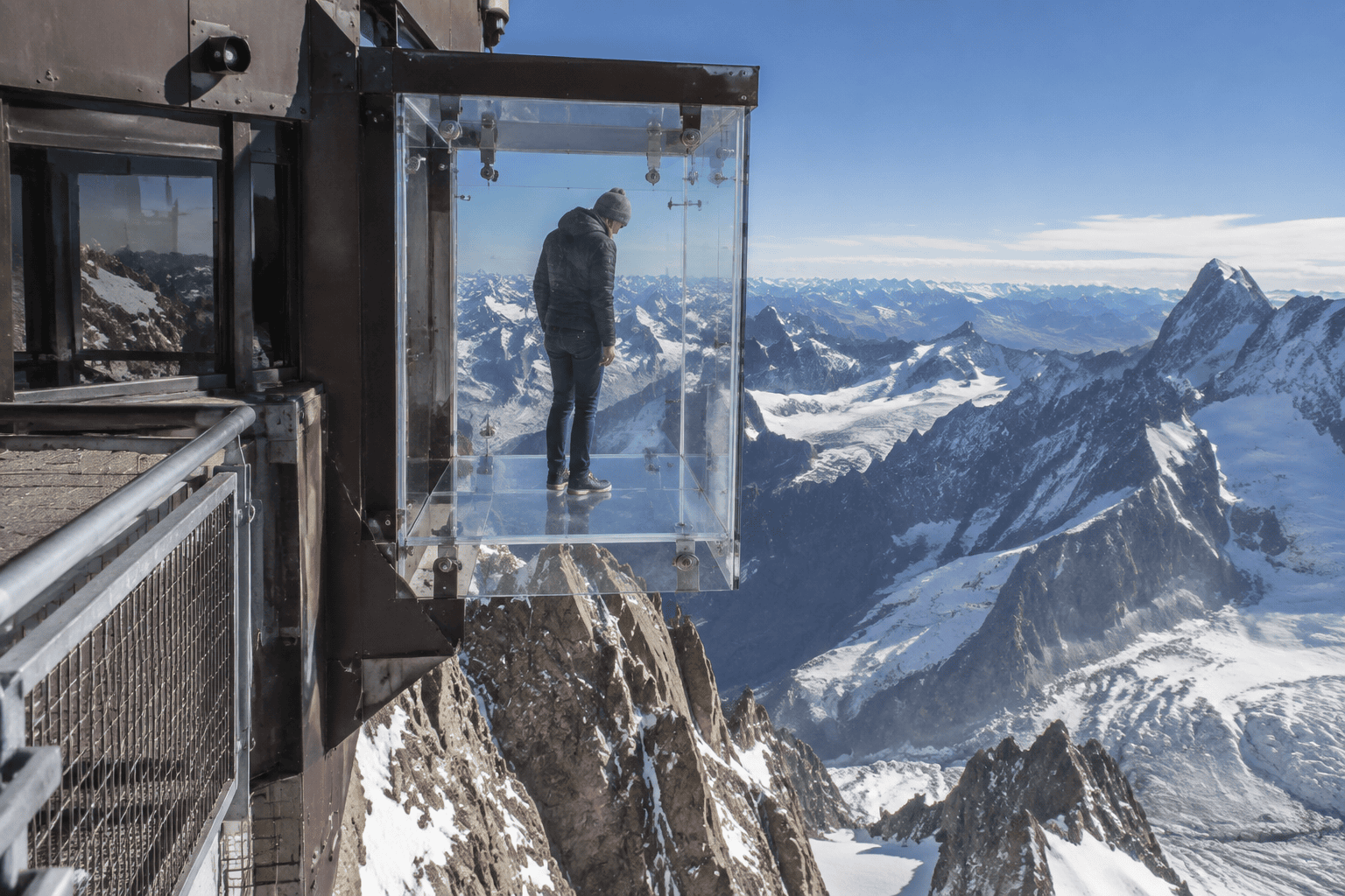 Une personne se tient dans une boîte vitrée suspendue à la station de l’Aiguille du Midi, face aux sommets enneigés.