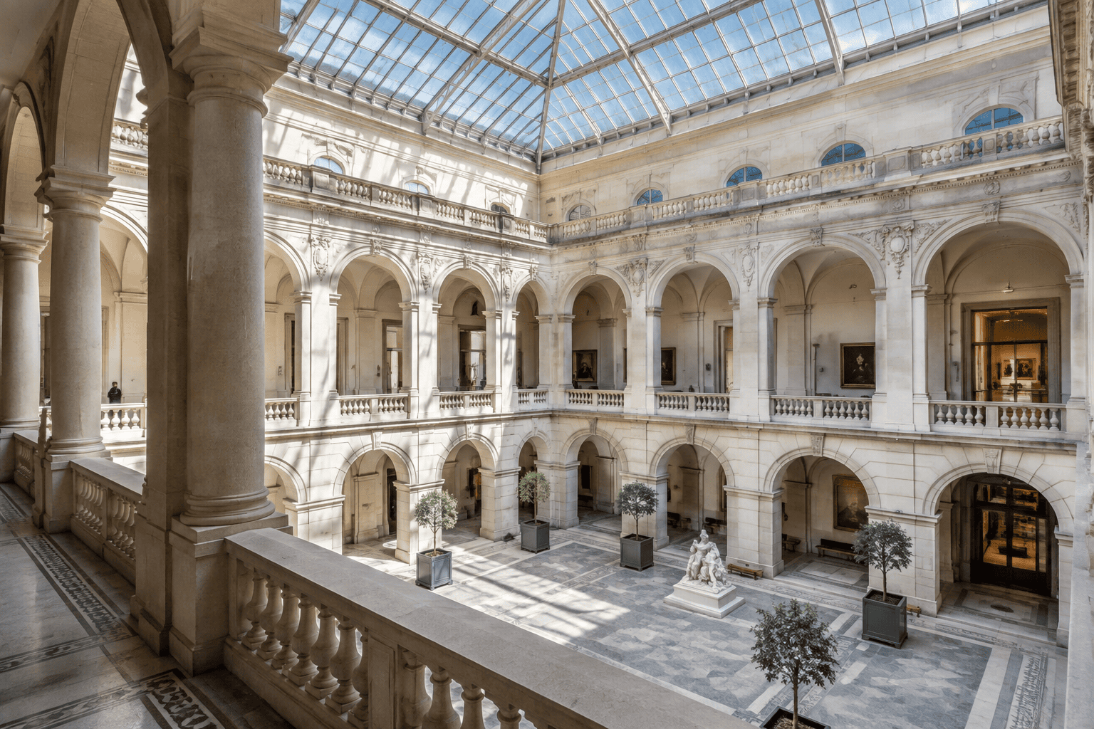 Cour intérieure à deux niveaux entourée de galeries en arcades et couverte d’une verrière, avec des arbres en pots et une sculpture au centre.