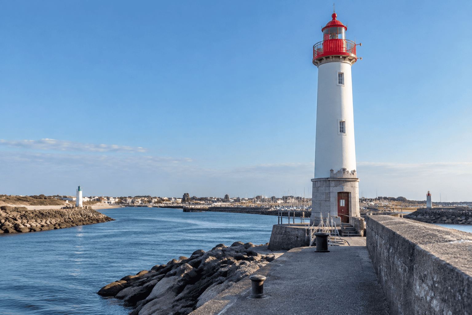 Un phare blanc surmonté d’une lanterne rouge se dresse au bout d’une digue, face à un chenal bordé de rochers.