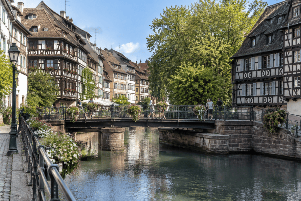 Une passerelle piétonne traverse un canal entre des maisons à colombages et des quais fleuris.