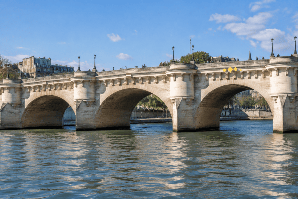 Un pont parisien en pierre aux arches arrondies traverse la Seine, avec l’eau au premier plan.
