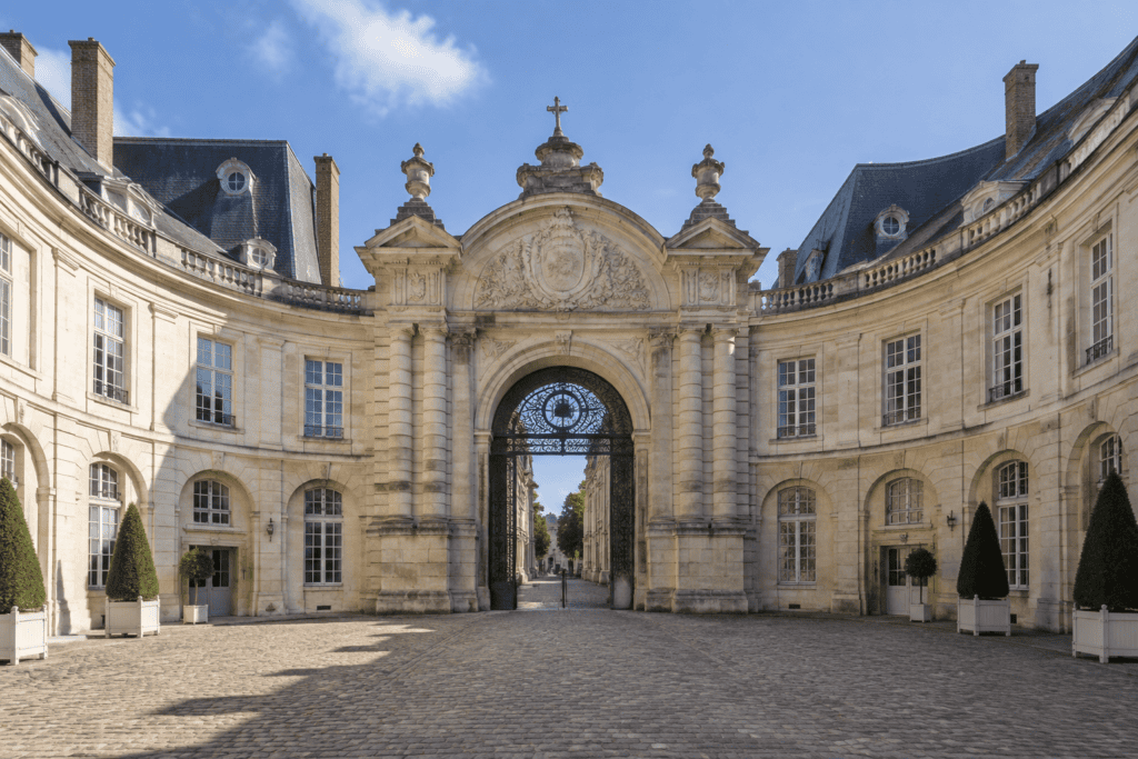 Portail sculpté au centre d’une cour bordée de façades incurvées, avec une grille en ferronnerie sous l’arche.