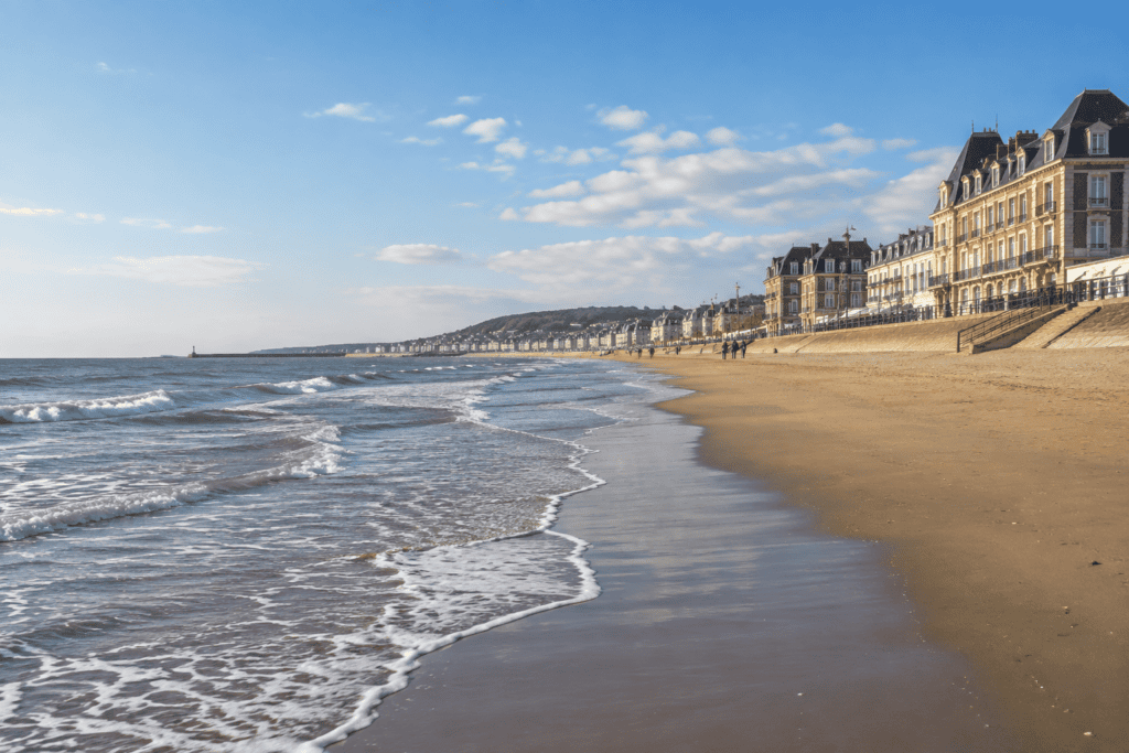 Le rivage de sable de Trouville-sur-Mer longe la mer, avec des vagues sur la gauche et des bâtiments en bord de plage à droite.
