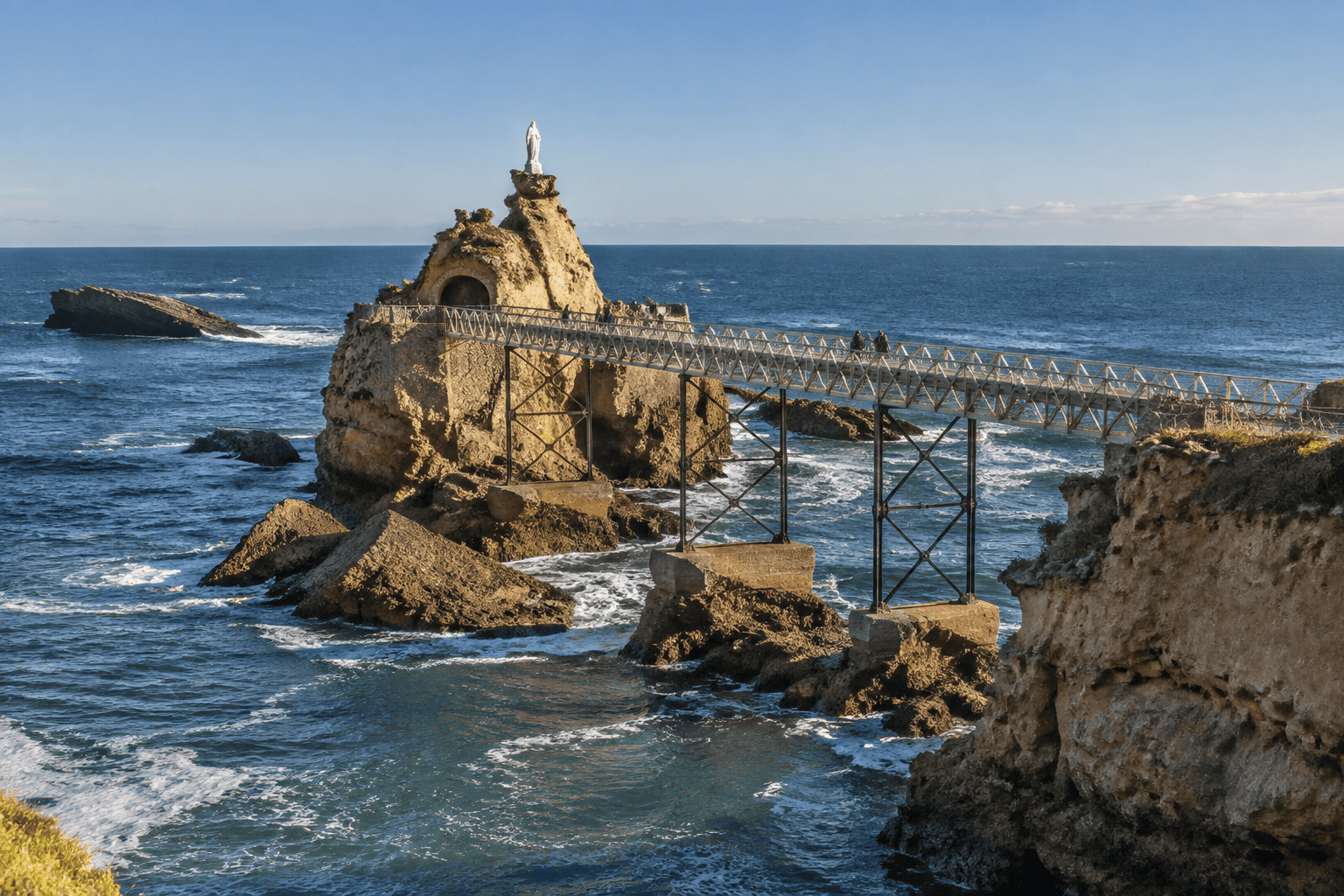 La passerelle métallique rejoint le rocher surmonté d’une statue blanche, au milieu des vagues de l’Atlantique.