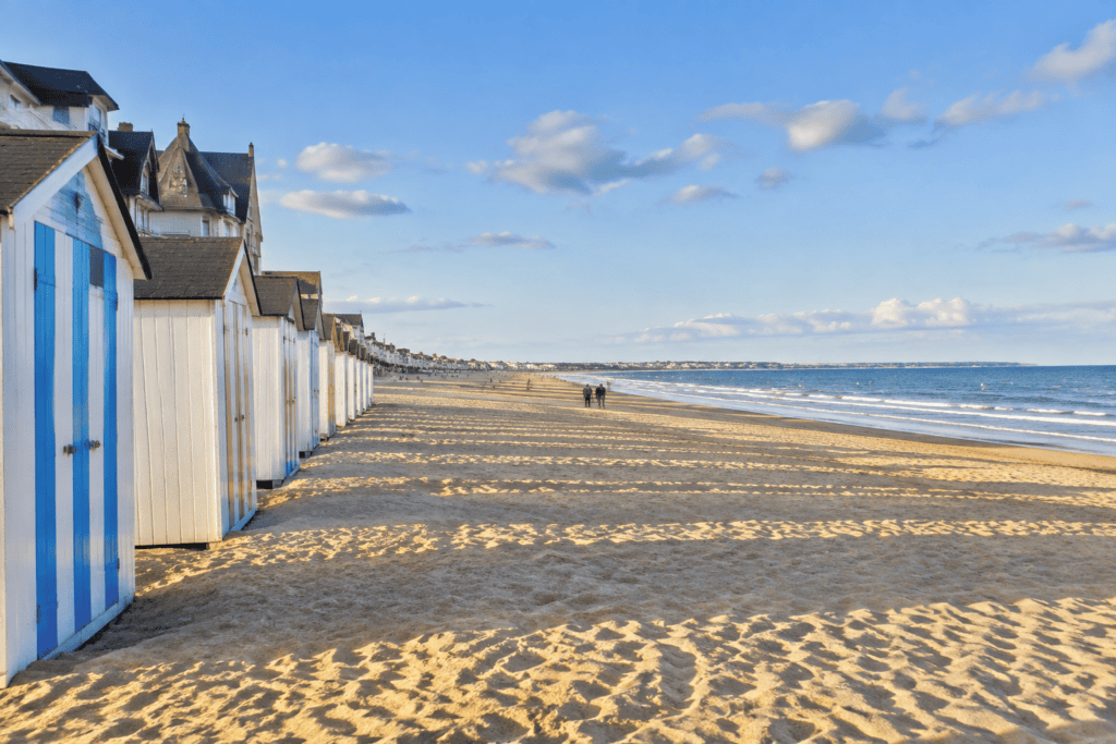 Des cabines blanches, dont certaines à portes bleues, bordent une plage de sable face à la mer.