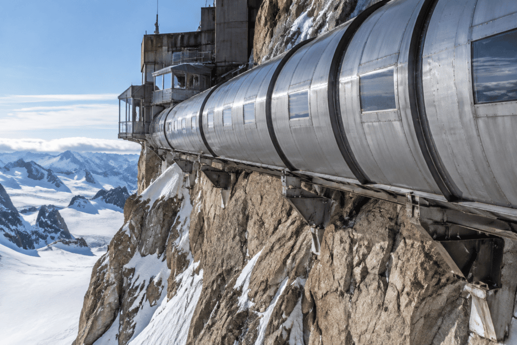 Une galerie métallique fermée longe le piton rocheux de l’Aiguille du Midi, au-dessus de pentes enneigées.
