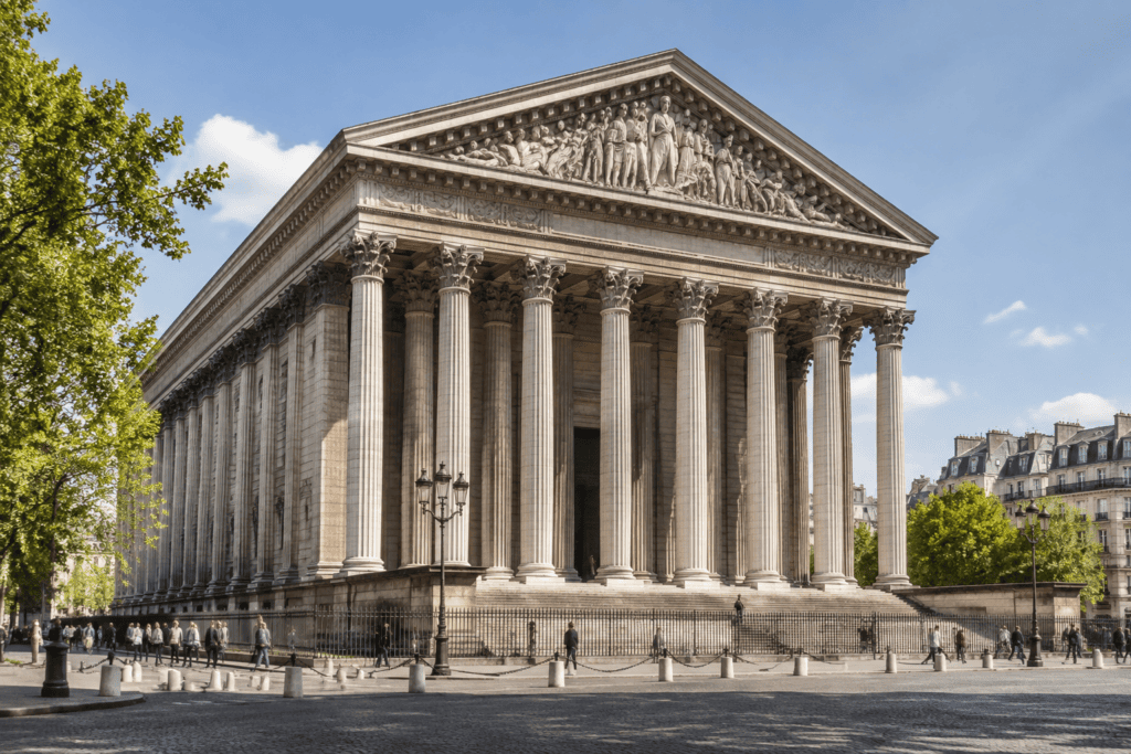 Façade à colonnes de l’église de la Madeleine, avec des arbres et des passants sur la place.