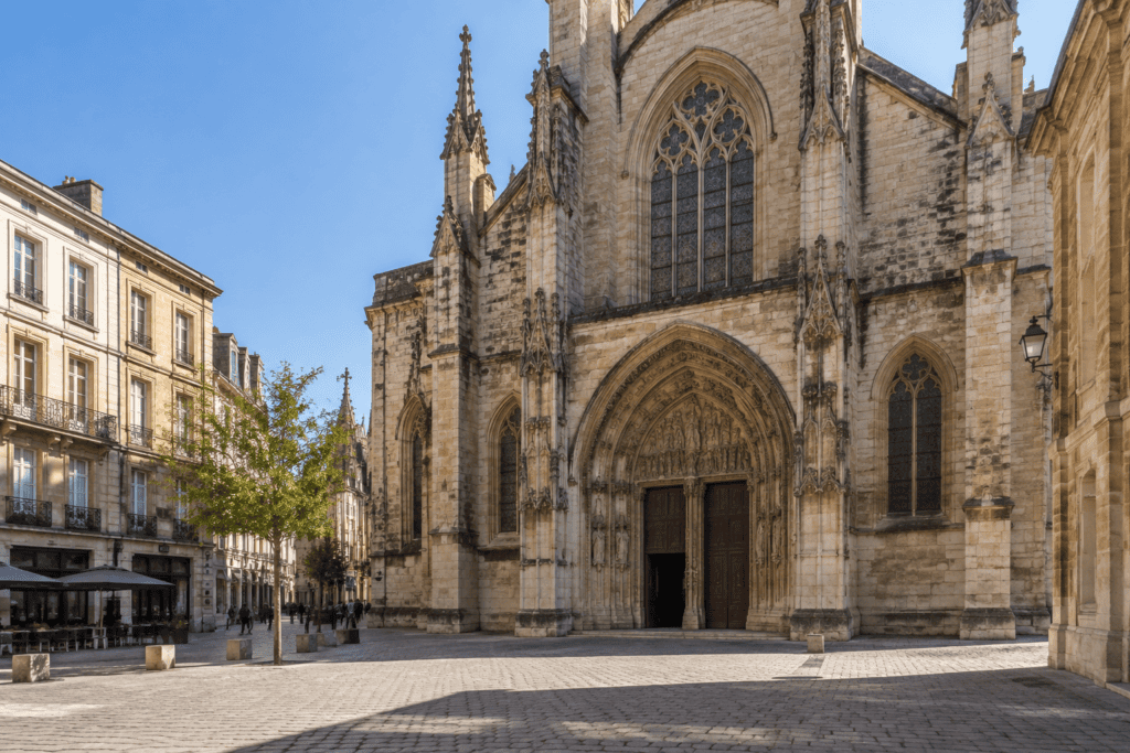 Façade en pierre d’une église gothique donnant sur une place pavée, avec des immeubles et des terrasses à proximité.