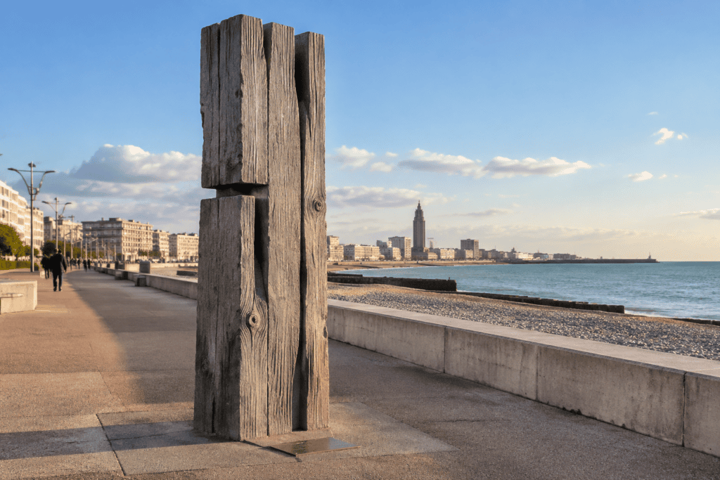Sculpture composée de hautes pièces de bois dressées au bord de la promenade, avec la plage et la mer à droite et les bâtiments du Havre à l’horizon.