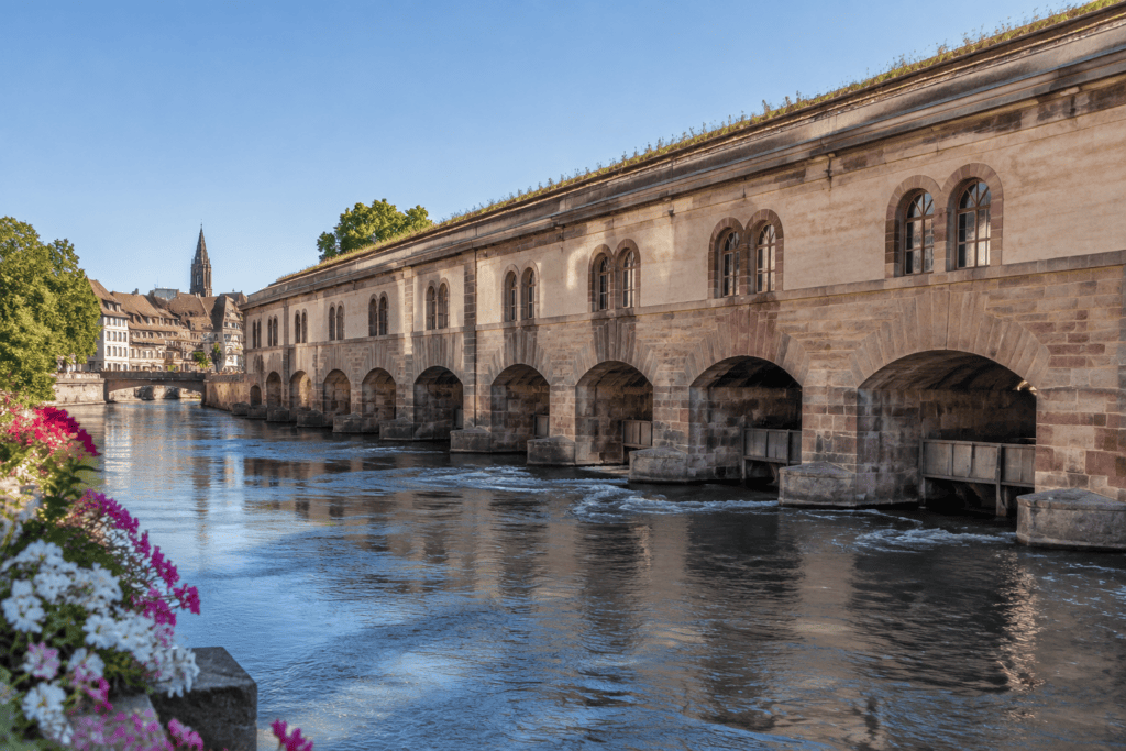 La façade de pierre du barrage Vauban et ses arches au-dessus de l’Ill, avec des bâtiments strasbourgeois visibles au loin.