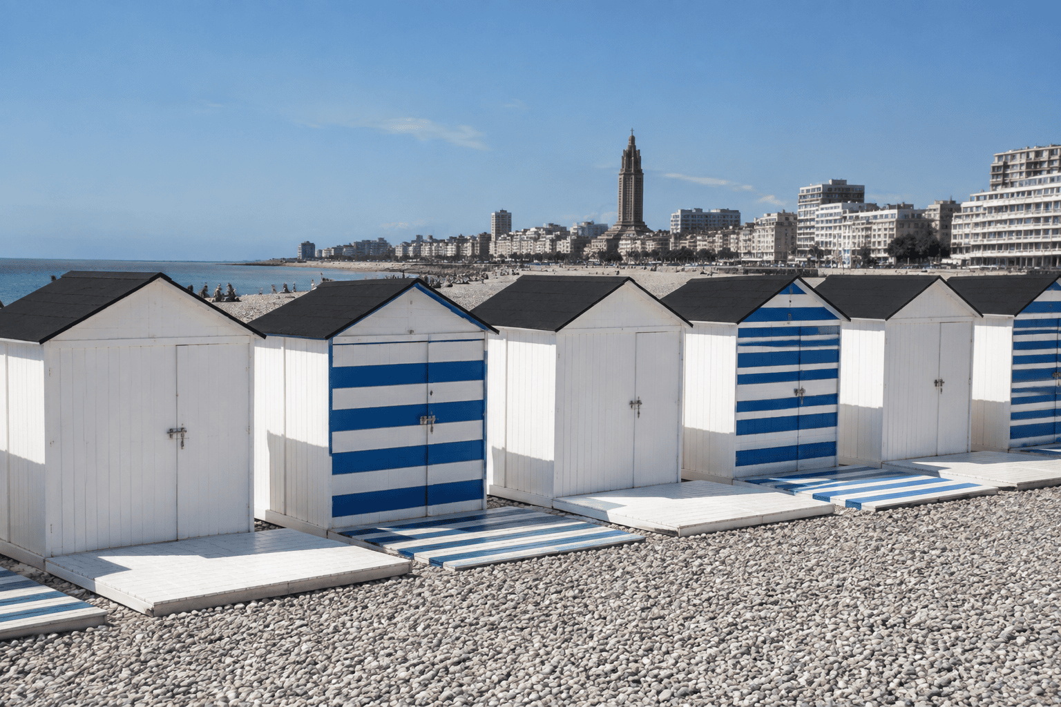 Rangée de cabanes blanches aux toits noirs, dont plusieurs présentent des rayures bleues, sur la plage de galets du Havre.