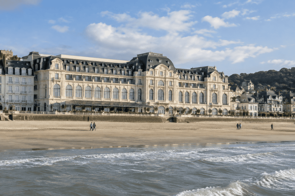 Longue façade d’hôtel en bord de mer, avec une plage de sable et des vagues au premier plan.