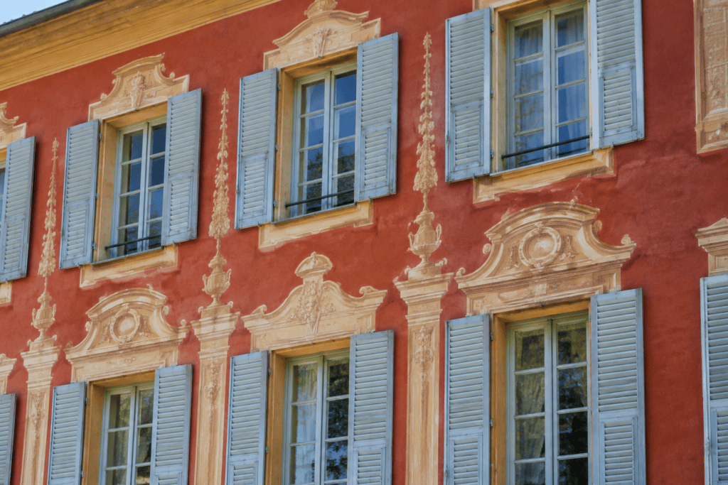 Façade rouge ornée de décors peints autour de fenêtres à volets bleus.
