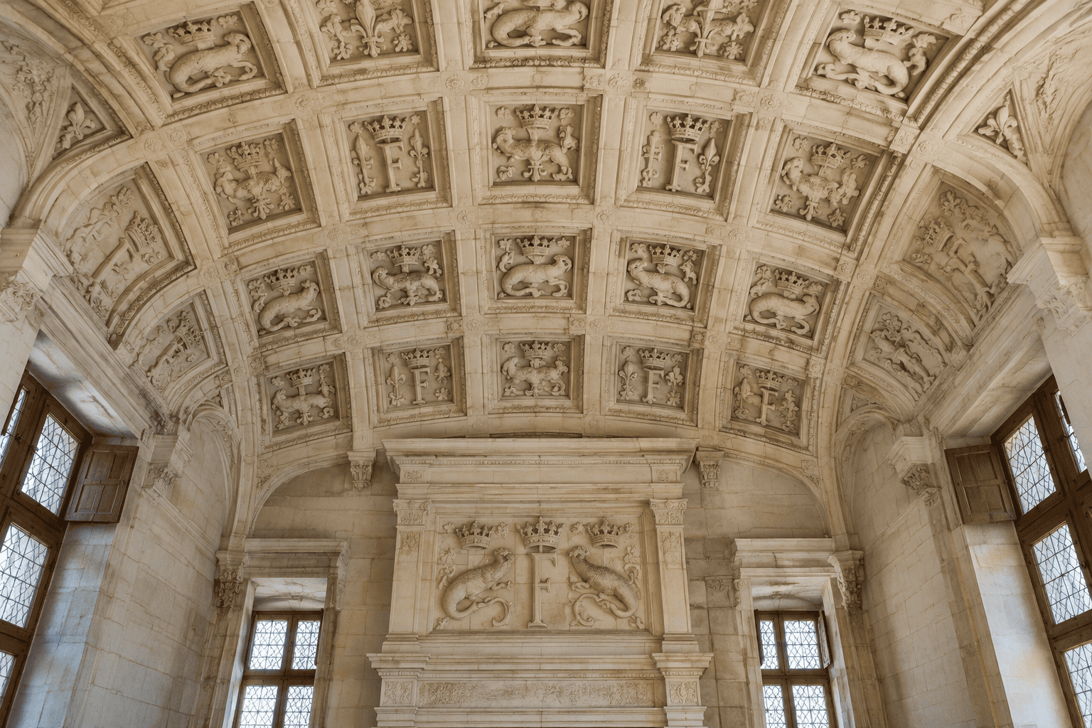 Voûte en pierre à caissons sculptés au-dessus d’une cheminée, dans une salle aux hautes fenêtres.