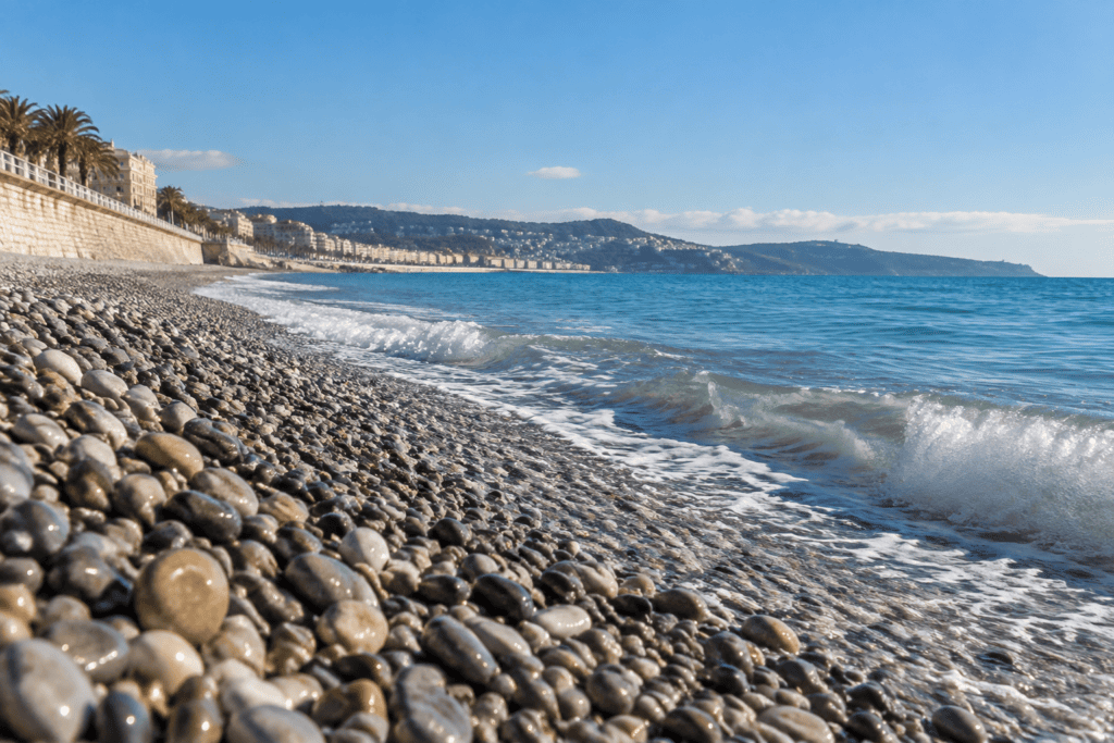 Galets au premier plan et vagues sur une plage de Nice, avec le front de mer et les collines en arrière-plan.