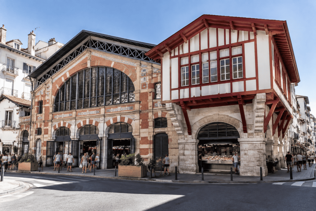 Deux halles de marché bordent une rue : l’une présente une façade de brique avec de grandes ouvertures cintrées, l’autre une structure blanche à colom