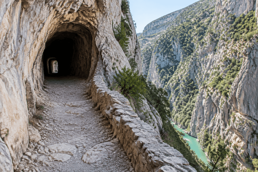 Un chemin de pierre traverse un tunnel creusé dans la roche, au-dessus d’une rivière turquoise encaissée entre des falaises.