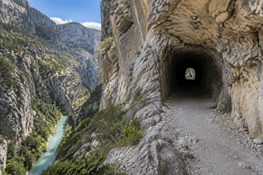 Un sentier rocheux mène à l’entrée sombre d’un tunnel creusé dans la falaise, au-dessus d’une rivière turquoise encaissée entre des parois calcaires.