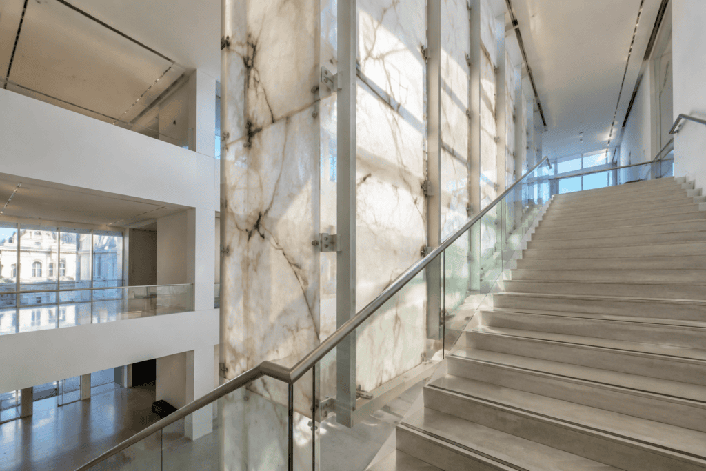 Un escalier longe une grande paroi translucide de marbre et de verre dans un espace intérieur contemporain.