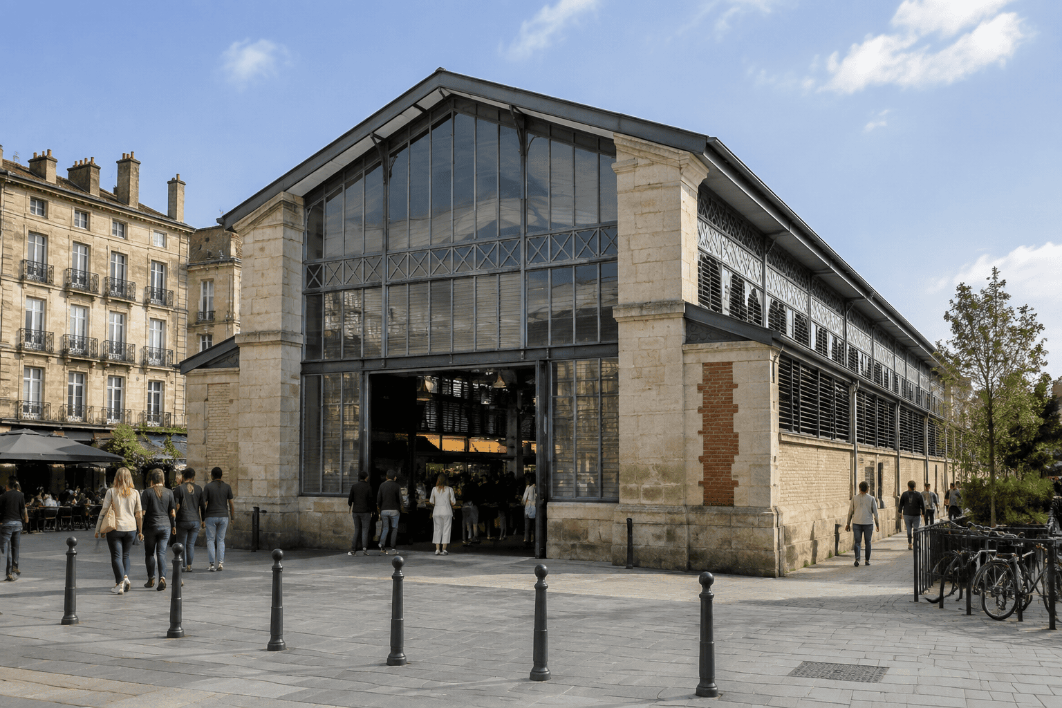 Entrée vitrée d’une halle de marché, avec des passants sur une place pavée.