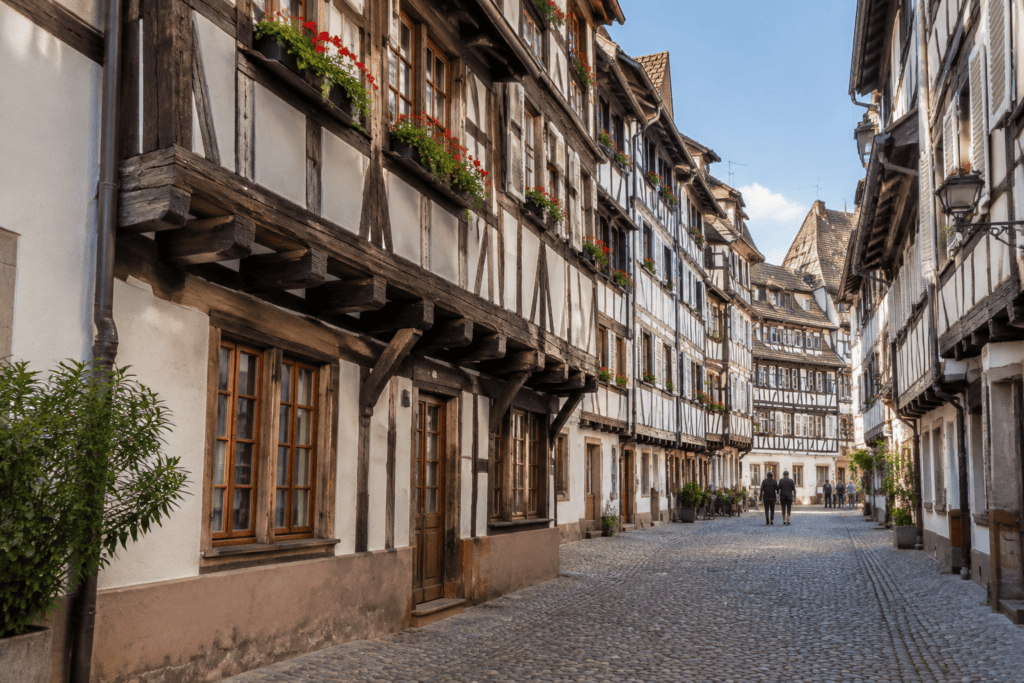 Ruelle pavée bordée de maisons à colombages aux étages en encorbellement, avec quelques passants au loin.