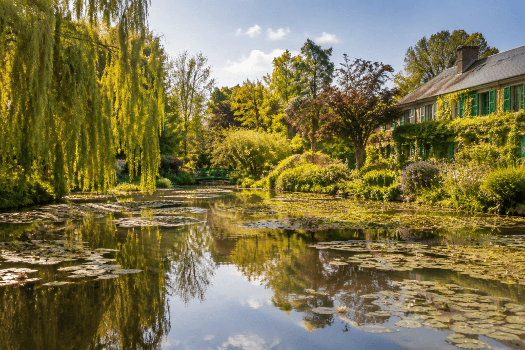 Bassin couvert de nénuphars, entouré d’arbres, avec une maison aux volets verts et un petit pont en arrière-plan.