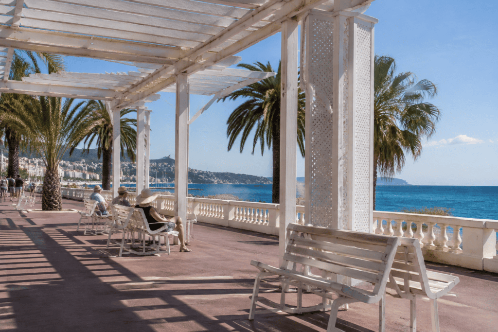 Des bancs sous une pergola blanche longent la mer, avec des palmiers et des promeneurs sur la promenade.