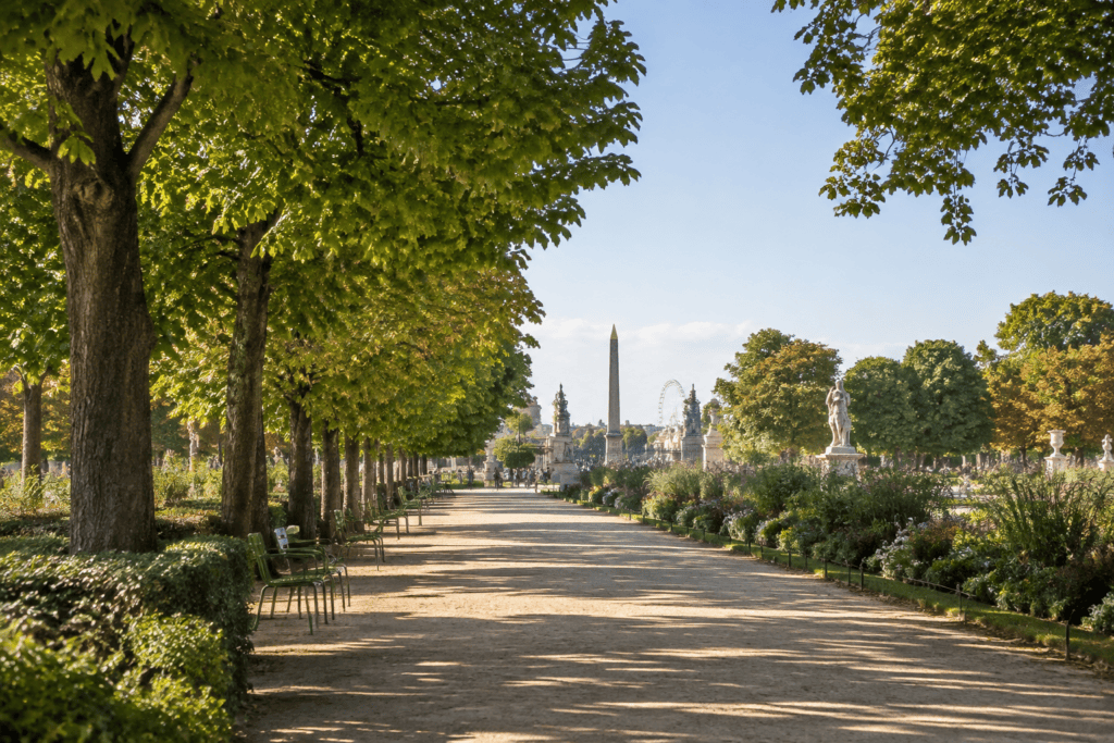 Allée en gravier bordée d’arbres, de massifs et de bancs, avec un obélisque visible au loin.