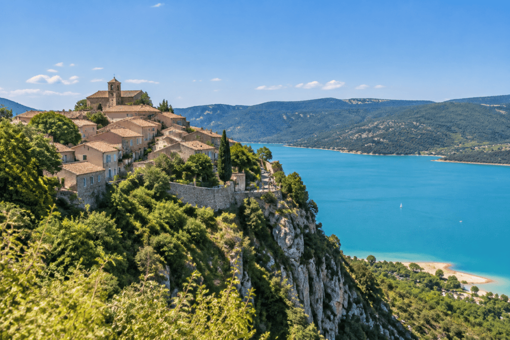 Maisons aux toits de tuiles regroupées sur un promontoire rocheux, avec le lac bleu et des collines à l’arrière-plan.