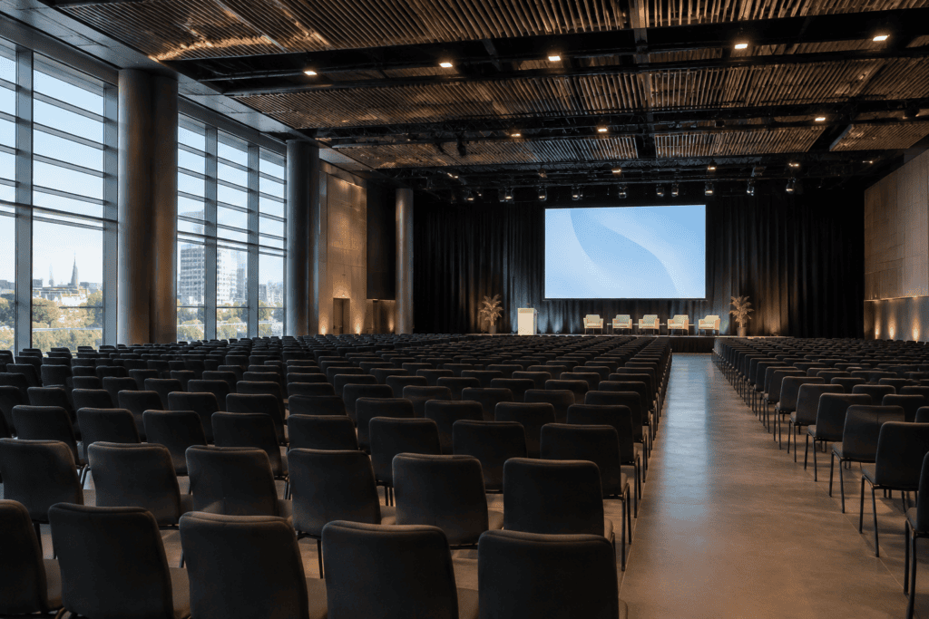 Rangées de chaises face à une scène avec un écran dans une grande salle de conférence.