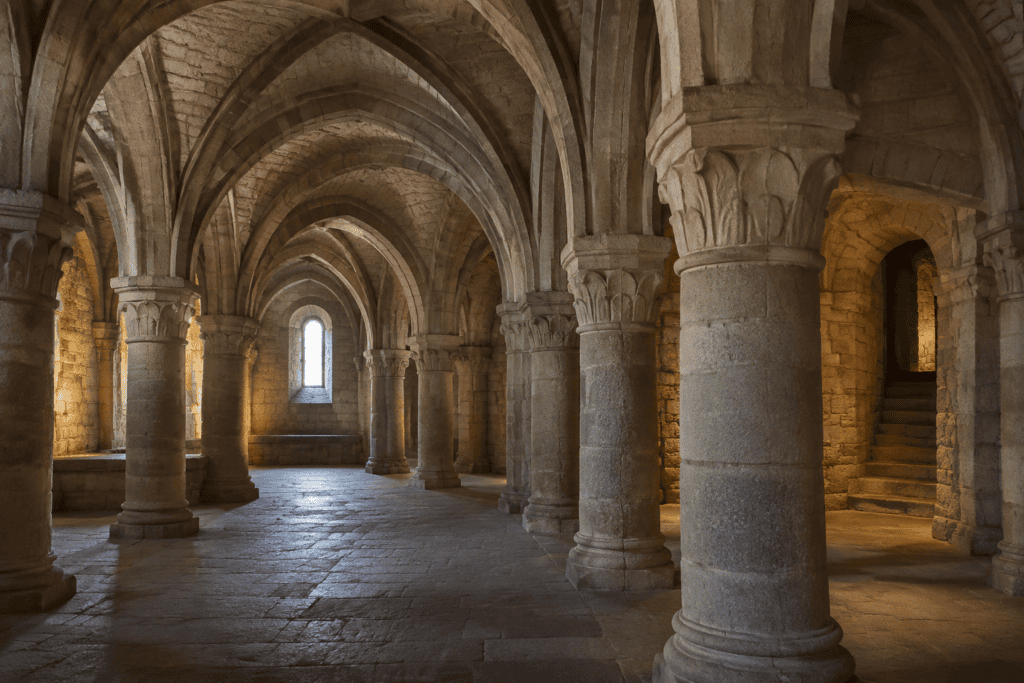 Intérieur voûté en pierre avec des piliers massifs, un sol dallé et une petite fenêtre.