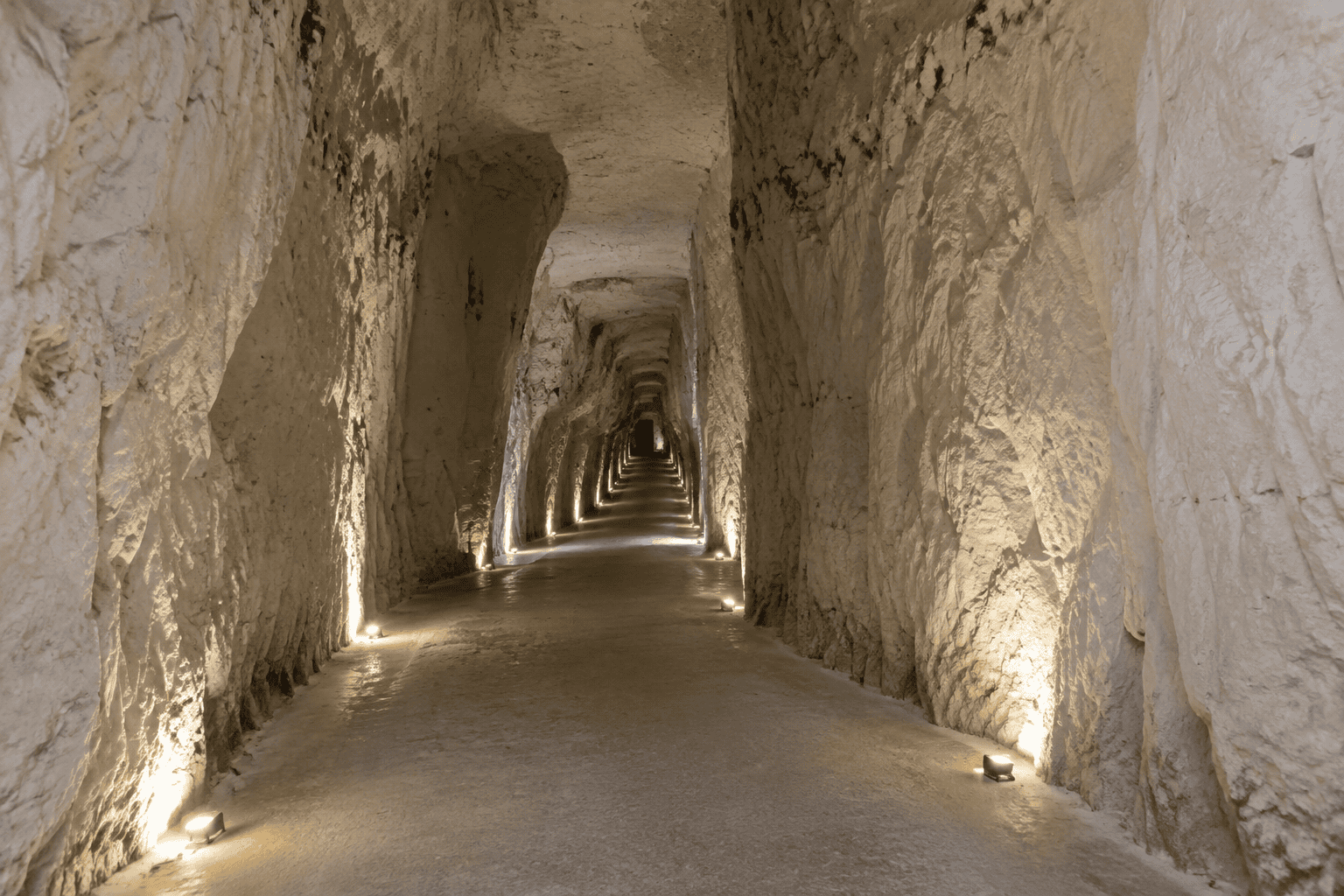 Un long passage souterrain aux parois claires et irrégulières, éclairé par des lampes au ras du sol.