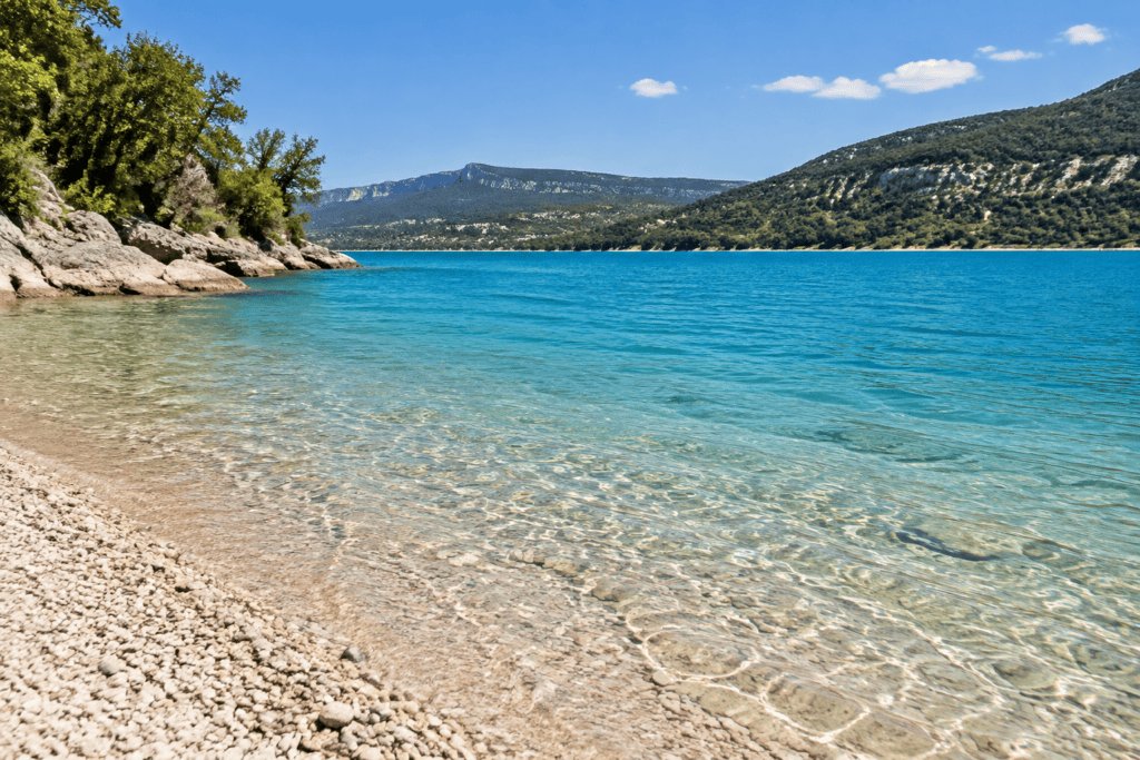 Une rive de galets descend dans une eau turquoise peu profonde, face à des collines boisées.