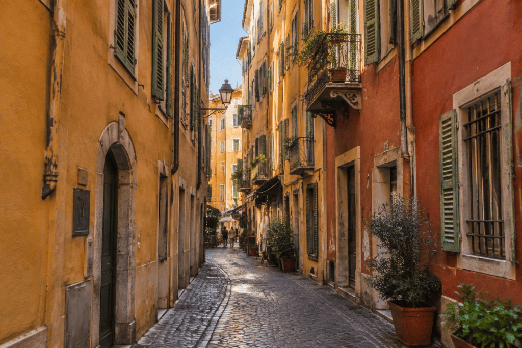 Une ruelle pavée bordée de façades ocre et rouges, avec volets persiennés et balcons en ferronnerie.