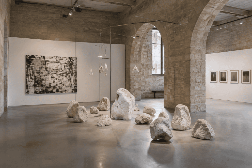 Des formes sculpturales claires sont disposées au sol dans une galerie aux murs de pierre, avec des œuvres accrochées aux murs.