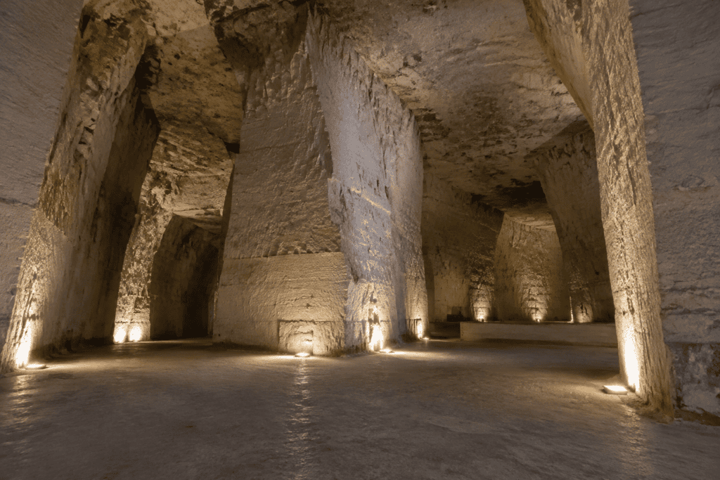 Salle souterraine aux hautes parois de craie, avec de larges piliers, un plafond irrégulier et un sol dégagé éclairé par des lampes basses.